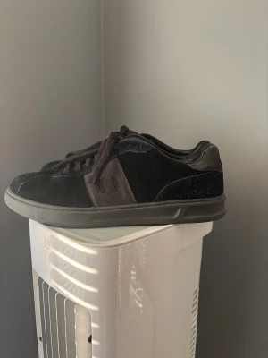 Svarta sneakers från Fred Perry mocka - Svarta sneakers från Fred Perry i mocka och skinn med klassisk laurel-wreath logga på sidan och hälen. Snygg, låg profil med rund tå och snörning. Sulan är svart och ovandelen har en mix av slät och ruggad mocka för en cool kontrast.