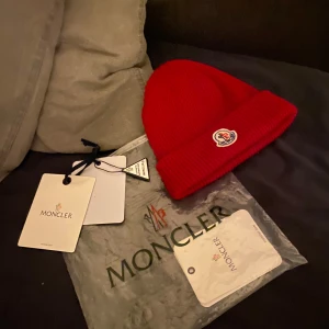 Röd Moncler mössa med logga - Snygg röd ribbstickad mössa från Moncler med klassisk uppvikt kant och ikonisk logopatch framtill. Perfekt för dig som vill ha en clean och lyxig look i vinter. Kommer med originalförpackning och tags.