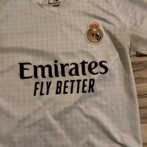 Real Madrid Bellingham fotbollströja - Säljer en vit Real Madrid fotbollströja med Bellingham och nummer 5 på ryggen. Tröjan har korta ärmar, klubbmärke på bröstet och Emirates Fly Better som sponsortryck. Materialet är lätt och andas, perfekt för match eller träning. Snyggt subtilt mönster över hela tröjan.