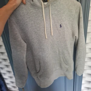Grå hoodie från Polo Ralph Lauren - Grå hoodie från Polo Ralph Lauren med klassisk broderad logga i mörkblått på bröstet. Tröjan har huva med vita snören, känguruficka och långa ärmar. Mjuk bomullskvalitet och relaxed passform, perfekt för chill dagar.