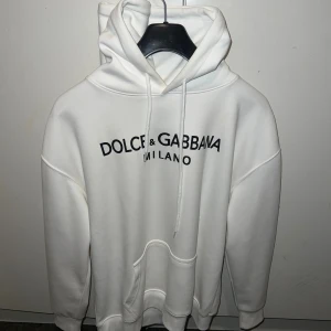 Vit hoodie Dolce & Gabbana Milano - Vit hoodie från Dolce & Gabbana med svart tryck 'Dolce & Gabbana Milano' på bröstet. Tröjan har huva med snörning, stor känguruficka och ribbade muddar. Perfekt för dig som gillar stilrena och exklusiva märken.