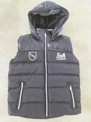 Svart dunväst med huva från SRK - Svart dunväst från SRK Basics med huva och silverfärgade dragkedjor. Västen har broderade loggor på bröstet, quiltad design och två fickor med dragkedja. Ribbad krage med beige och vit rand. Perfekt för dig som vill ha en cool och sportig look.