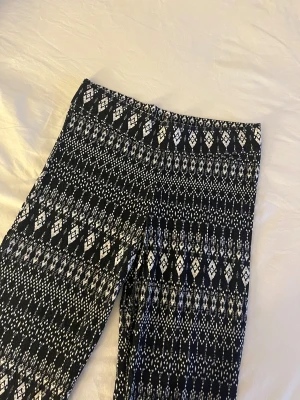 Svarta mönstrade bootcut byxor - Säljer ett par svarta bootcut byxor med vitt grafiskt mönster. Byxorna har mellan midja och är tillverkade i ett mjukt, stretchigt material som sitter skönt. Perfekta för dig som gillar att sticka ut med coola mönster och bekväm passform.