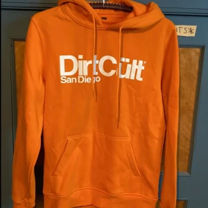 Orange hoodie från DirtCult - Säljer en orange hoodie från DirtCult med vit logga och texten 'San Diego' på bröstet. Tröjan har huva med snörning, magficka och långa ärmar. Perfekt för dig som gillar streetwear och vill sticka ut med färg.