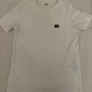 Vit t-shirt från C.P. Company XL - Vit t-shirt från C.P. Company i storlek XL. Klassisk design med rund hals och korta ärmar, samt en liten svart patch med logga på bröstet. Tillverkad i mjuk bomull som känns skön mot huden. Perfekt för en clean och stilren look.