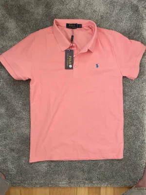  Ralph Lauren piké - Snygg rosa pikétröja från Polo Ralph Lauren med klassisk krage och korta ärmar. Tröjan har blå broderad logga på bröstet och knäppning framtill. Perfekt för en chill och stilren look.