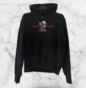 Svart Helmut Lang hoodie med tryck - Svart hoodie från Helmut Lang med stort grafiskt tryck och röd logga på bröstet. Tröjan har huva med snörning, ribbade muddar och en klassisk känguruficka framtill. Perfekt för dig som gillar streetwear och vill sticka ut med en cool design. Tyvärr har Hoodien gått lite sönder på en av repen men inget man stör sig på. Ny pris ca :2000, kan diskuteras vid snabb affär