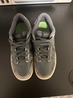 Nike Dunk Low grå/svart med lime sula - Nike Dunk Low sneakers i mörkgrå och svart med slitna detaljer och limegröna sulor. Skorna har snörning, perforerad tå och klassisk Nike-logga på hälen. Ovandelen är i syntet och skinn, med låg profil och platt sula. Perfekt för dig som gillar streetwear och unik stil.