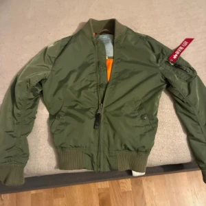 Grön bomberjacka Alpha Industries XS - Säljer en klassisk grön bomberjacka från Alpha Industries i storlek XS. Jackan har orange foder, ribbade muddar och den ikoniska röda 'Remove Before Flight'-taggen på ärmen. Tillverkad i slitstark polyester och stängs med dragkedja.