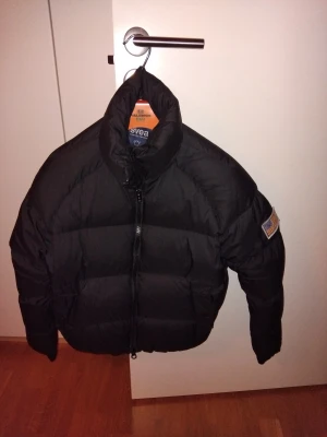 Svart dun pufferjacka från Svea - Svart dun pufferjacka från Svea med hög krage och dragkedja framtill. Jackan har en patch med amerikansk flagga på ena ärmen och en klassisk, vadderad look som håller dig varm under kalla dagar.
