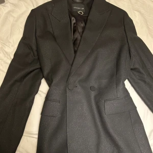 Svart glittrig kostym från Stockholm LM - Snygg svart kostym från Stockholm LM med diskret glittrigt tyg. Kavajen har klassisk krage, två knappar och fickor med lock. Byxorna är raka och har hällor för bälte. Perfekt för dig som vill sticka ut med stilren glittereffekt. Kavaj i storlek M och byxa i storlek 42