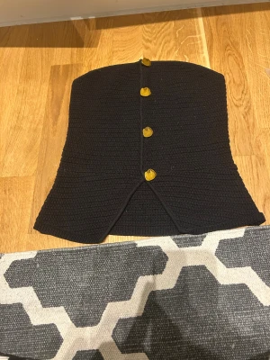 Svart stickad tubtopp med knappar - Säljer en svart stickad tubtopp med snygga guldfärgade knappar framtill. Toppen har en bandeauform och en liten slits nedtill som ger extra edge. Perfekt för dig som vill ha en unik och trendig topp till din outfit.