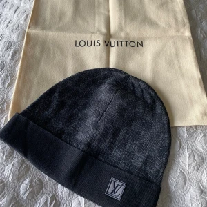 LV Mössa Svart - Exklusiv svart mössa från LV med diskret rutigt mönster och klassisk LV-logga framtill. Mössan har uppvikt kant och är tillverkad i mjukt material som känns lyxigt mot huden. Perfekt accessoar för att lyfta din vinterstil.