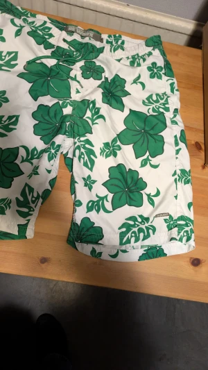 Blommiga badshorts från Blend - Snygga vita badshorts från Blend med stora gröna blommor och bladmönster. De har grön snörning i midjan och meshfoder på insidan. Perfekta för strandhäng eller poolparty, med en chill vibe och tropisk känsla.