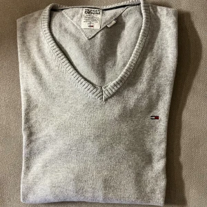 Tommy Hilfiger tröja grå - Snygg grå v-ringad tröja från Tommy Hilfiger Denim med diskret logga broderad på bröstet. Långärmad tröja. Tröjan har ribbad kant vid halsen och är tillverkad i mjuk bomull, perfekt för en clean och stilren look.