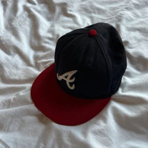 Atlanta Braves keps från New Era - Snygg Atlanta Braves 59FIFTY keps från New Era med svart krona, röd skärm och röd knapp på toppen. Broderad vit Braves-logga på framsidan. Klassisk baseballstil, perfekt för dig som gillar sportig streetwear.