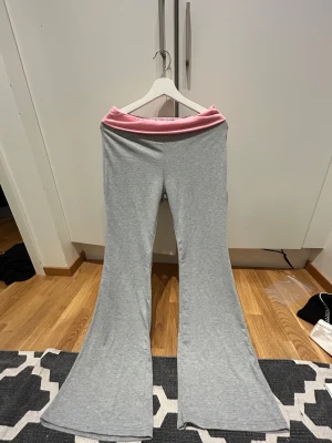 Grå bootcut mjukisbyxor med rosa midja - Supermjuka grå mjukisbyxor med bred bootcut och en snygg rosa midja. Perfekta för chill dagar eller när du vill ha något bekvämt men ändå stilrent. Materialet är stretchigt och skönt mot huden.