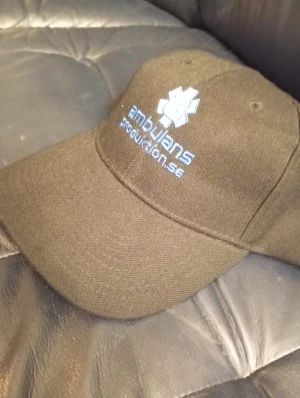 Svart keps Ambulansproduktion.se - Svart keps med böjd skärm och broderad logga från Ambulansproduktion.se i blått och vitt framtill. Klassisk modell med ventilerande hål upptill, tillverkad i bomull. Perfekt för dig som vill ha en clean och enkel look med lite edge.