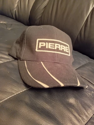 Svart keps från Pierre med vita detaljer - Svart keps från Pierre med broderad logga framtill i vitt. Skärmen har coola vita linjer som ger en sportig vibe. Klassisk form med böjd skärm och ventilerande hål upptill. Perfekt för dig som gillar enkel och stilren streetwear.