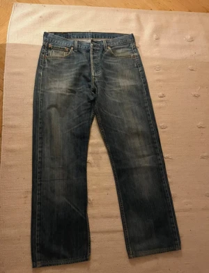 Levi's 501 blå jeans W34 L36 - Klassiska Levi's 501 jeans i blå tvätt med raka ben och fem fickor. Jeansen har en normal passform och är tillverkade i slitstarkt bomullsdenim. Ikonisk läderpatch bak i midjan och knappgylf. Perfekta för dig som gillar tidlös stil. O.B.S att dom sitter ungefär spm 32,32 istället 