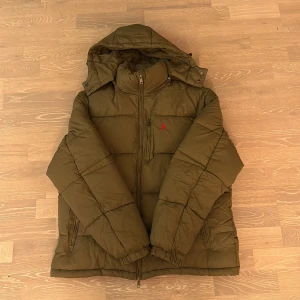 Olivgrön pufferjacka från Polo Ralph Lauren - Olivgrön pufferjacka från Polo Ralph Lauren med huva, dragkedja och bröstficka. Jackan har det klassiska röda Polo-broderiet på bröstet och är tillverkad i syntetmaterial. Perfekt för kalla dagar med sin vadderade design och snygga look.