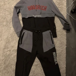 Svart hoodie och grå mjukisbyxor från Hoodrich - Snygga mjukisbyxor och hoodie från Hoodrich i svart och grått med logga tryckt på benet och bröstet. Byxorna har snörning i midjan och dragkedjor på fickorna för extra stil och funktion. Perfekta för chill eller streetwear-look. Byxor strlk L och hoodie i strlk M