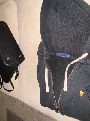 Svart hoodie från Polo Ralph Lauren - Svart hoodie från Polo Ralph Lauren med gul broderad logga på bröstet. Tröjan har huva med vita snören, dragkedja framtill och mjukt fleecefoder. Perfekt för chill dagar och har en klassisk streetstil. Storlek S.