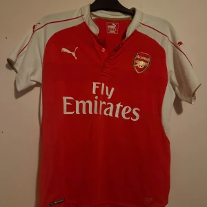 Arsenal Puma Santi Cazorla tröja - Röd och vit Arsenal fotbollströja från Puma med Santi Cazorla och nummer 19 på ryggen. Klassisk design med klubbmärke, Fly Emirates-tryck och korta ärmar. Perfekt för dig som älskar Premier League och vill visa ditt stöd för Arsenal. Tröjan är köpt i shoppen vid Arsenals arena.