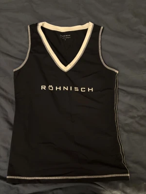 Träningströja - Svart träningslinne från Röhnisch med vit kontrastkant och v-ringad hals. Toppen är ärmlös och har en sportig look med logotypen tryckt framtill. Materialet är mjukt och stretchigt, perfekt för aktiva dagar eller träning. Storlek xs ny utan prislapp. Nypris 900kr