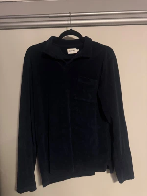 Mörkblå frote långärmad piké från East West - Snygg mörkblå frote långärmad frote från East West . Tröjan har klassisk krage, lång ärm och en bröstficka som ger en chill vibe. Perfekt för dig som gillar stilrena och bekväma plagg med lite retrokänsla.