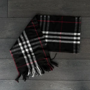 Svart Burberry halsduk - Stilren svart halsduk med klassiskt rutigt mönster i vitt, grått och tunna röda linjer. Mjukt material och snygga fransar i ändarna ger en cool vibe. Perfekt att slänga över axlarna när det är kyligt ute.