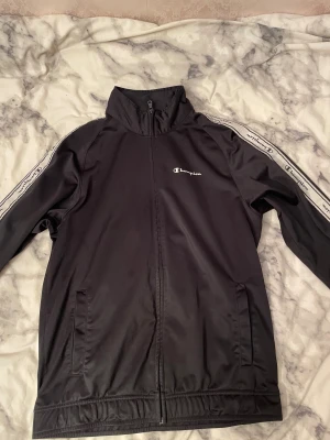 Svart vindjacka från Champion M - Svart vindjacka från Champion i storlek medium. Jackan har dragkedja framtill, höga krage och vita logoband längs ärmarna med Champion-tryck. Tillverkad i polyester, perfekt för sportiga dagar. Snygg och enkel design med fickor på sidorna.