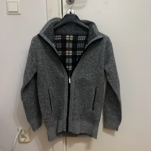 Verani Zip-Up storlek S - Mysig grå stickad kofta med hel dragkedja och höga ribbade muddar. Insidan är fodrad med rutigt tyg i blått, vitt och brunt. Hög krage för extra värme. Storlek S, originalpris är 799kr från Verani. Säljer den för att jag fick fel tröja när jag beställde. Aldrig använt den. Tröjan saknar fickor
