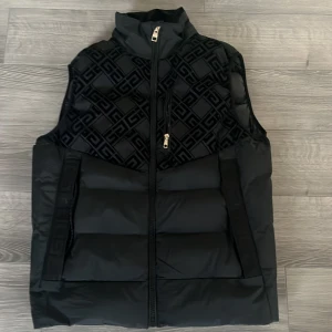 Svart dunväst från Glorious Gangsta - Svart dunväst med hög krage och dragkedja framtill från Glorious Gangsta. Övre delen har ett snyggt geometriskt mönster i svart, medan resten är quiltad och puffig. Västen har två fickor och detaljer i guld på dragkedjan. Perfekt för att addera edge till din outfit.
