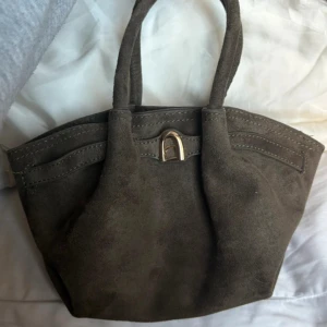 Brun mocka handväska med gulddetalj - Snygg handväska i brun mocka med mjuk struktur och två rejäla handtag. Väskan har en unik guldfärgad metallspänne framtill och rymligt svart innerfoder. Perfekt storlek för att få plats med allt du behöver och ger en cool vibe till din outfit.