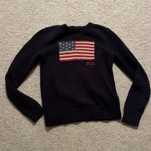 Mörkblå stickad tröja Polo Ralph Lauren - Snygg mörkblå stickad tröja från Polo Ralph Lauren med amerikansk flagga broderad på bröstet och RL-detalj i rött. Tröjan har långärmad passform, ribbade muddar och rund hals. Perfekt för dig som gillar klassisk stil med en twist. Tröjan är äkta och ser ut som ny! 