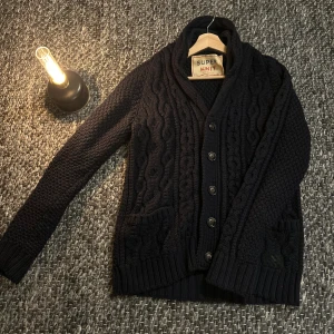 Cardigan - Säljer en svart, grovstickad kofta med snyggt kabelmönster och ribbade muddar. Koftan har knappar framtill, två fickor och en skön krage som ger extra värme. Perfekt för dig som gillar klassisk stil med en modern twist.