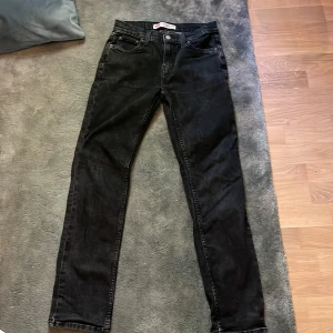 Svarta Levi's 502 jeans taper - Svarta jeans från Levi's, modell 502 Regular Taper. Snygg, klassisk femficksdesign med smalare ben och normal passform. Jeansen har en mörk tvätt och är tillverkade i slitstarkt denim med knapp och dragkedja framtill. Perfekt för dig som gillar stilrena och tidlösa jeans.