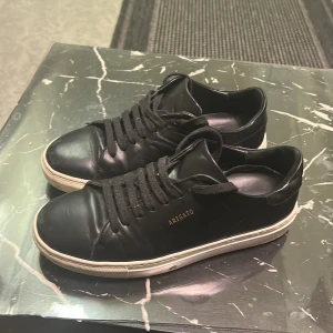 Svarta sneakers från Axel Arigato - Snygga svarta sneakers från Axel Arigato med klassisk låg profil och vit platt sula. Skorna har svart snörning och är tillverkade i slätt skinn med guldfärgad logga på sidan. Perfekta för dig som gillar minimalistisk och stilren design. Köpta för 1650kr
