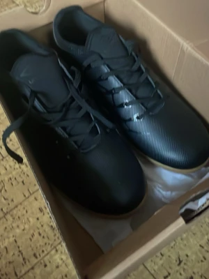 Svarta fotbollsskor från Umbro - Säljer ett par svarta fotbollsskor från Umbro med snygga detaljer och snörning. Skorna har en låg profil och platt sula som passar perfekt för inomhusspel. Materialet är syntet och designen är stilren med diskreta mönster längs sidorna.