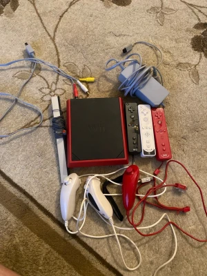 Nintendo Wii Mini - Nintendo Wii Mini med tre Wii-fjärrkontroller (svart, vit, röd), tre nunchuks (två vita, en röd), strömadapter, AV-kabel och sensorbar. Konsolen och tillbehören ser ut att vara i gott skick med mindre yttre slitage. Perfekt för familjespel och partyspel.