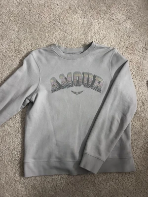 Grå sweatshirt från Zadig & Voltaire - Snygg grå sweatshirt från Zadig & Voltaire med texten 'AMOUR' i glittriga stenar på bröstet. Tröjan har rund hals, långa ärmar och en relaxed passform. Perfekt att matcha med jeans för en cool och avslappnad look.