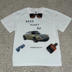 Vit t-shirt med Porsche 911 print - Snygg vit t-shirt i bomull med tryck på bröstet som visar en grön Porsche 911 och texten 'Need money for Porsche 911'. Klassisk rund hals och korta ärmar. Perfekt för dig som gillar bilar och vill sticka ut med en cool statement-tee.
