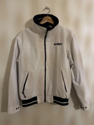 Vit vindjacka från RCMN - Snygg vit vindjacka från RCMN med svart dragkedja och detaljer. Jackan har hög krage, två fickor med svarta kanter och muddar med svarta och vita ränder. Perfekt för dig som gillar sportig stil och vill ha något lätt och fräscht.