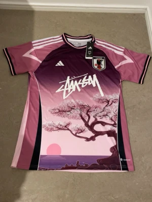 Adidas Japan x Stüssy fotbollströja  - Säljer en limited edition Japan landslagströja från Adidas i samarbete med Stüssy. Tröjan har en snygg rosa och lila gradient, grafiskt körsbärsträd och solnedgång på framsidan, samt broderat Japan-märke. Tillverkad i lätt polyester med Aeroready-teknologi.