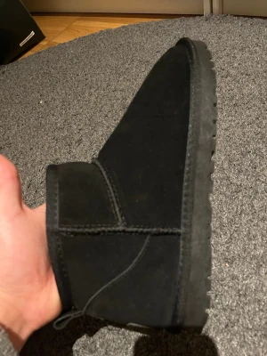 Svarta UGG boots i mocka - Säljer ett par svarta UGG boots med rund tå och platt sula. Skorna är tillverkade i mjuk mocka och har en fluffig insida som håller fötterna varma. Klassisk UGG-logga baktill och enkel design som passar till många stilar.