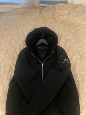 Vinter jacka  Moose Knuckles  - Svart pufferjacka från Moose Knuckles med stor huva och snyggt emblem på ärmen. Jackan har quiltad insida, dragkedja framtill och en riktigt schysst oversized look. Perfekt för kalla dagar och har en lyxig vibe med mjukt material.