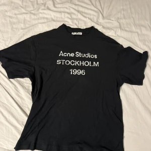 Svart Acne Studios t-shirt Stockholm - Svart t-shirt från Acne Studios med tryck i vitt på bröstet: 'Acne Studios STOCKHOLM 1996'. Klassisk rund hals och korta ärmar. Materialet känns som mjuk bomull, perfekt för en chill och stilren look. Köptes på Plick kvitto finns