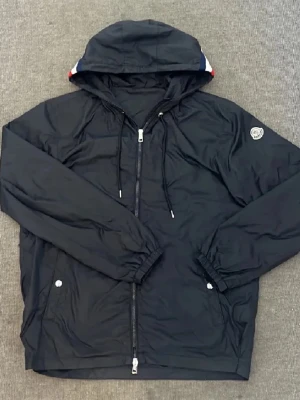 Svart vindjacka från Moncler - Svart vindjacka från Moncler med huva och snörning, hel dragkedja framtill och två fickor med tryckknappar. Jackan har Moncler-logga på ärmen och diskreta detaljer i rött, vitt och blått på huvan. Perfekt för dig som gillar stilrena och sportiga plagg. D ända är att dragkedjan gått sönder men den går ändå att knäppa och man ser ingeting att den e sönder 