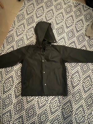 Svart regnjacka från Stutterheim - En näst intill oanvänd svart regnjacka från Stutterheim med huva och tryckknappar framtill. Jackan har två fickor framtill.  Ordinarie pris 1200kr, säljes för 700kr. Storlek 9-11 år